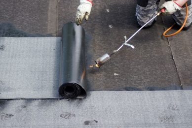 Pracownik przykleja papę termozgrzewalną do dachu za pomocą palnika gazowego.